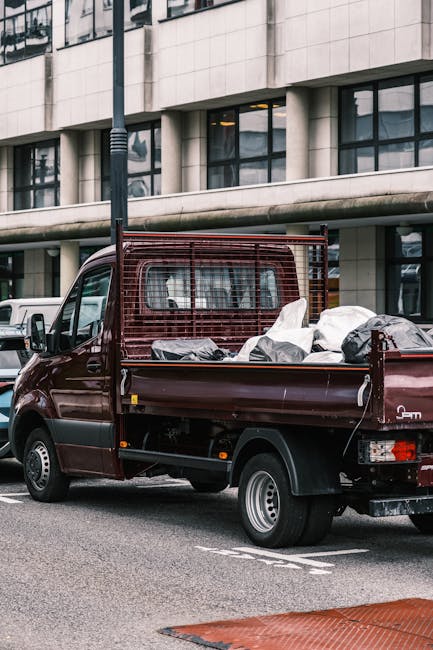 A small burgundy flatbed truck parked on an urban street in front of a modern office building with large glass windows. The truck's open cargo area is filled with various types of waste materials, including black and white plastic bags, some of which are loosely tied, and other mixed debris. The truck features a metal mesh barrier behind the cab, providing separation between the driver and the load, and the rear tailgate is down to accommodate the rubbish. The scene suggests a temporary collection of discarded items, potentially part of an on-site clearance or private waste collection service, supported by companies like House Clearance Kingston. The background includes other parked vehicles and a streetlamp, emphasizing the urban setting, while the natural daylight indicates a daytime operation. The overall environment reflects an organised yet informal rubbish removal process, typical in scenarios where independent collection or alternative waste handling methods are employed outside of municipal services.