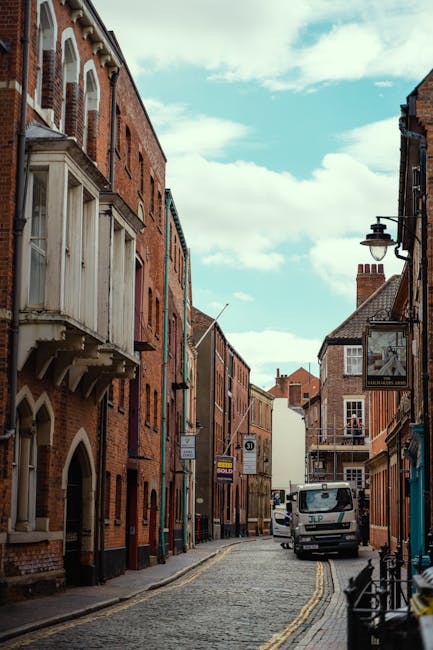 A narrow cobblestone street in Kingston lined with multi-story brick buildings featuring a mix of red and brown tones, irregular façade designs, and small bay windows. The buildings have various architectural details such as decorative arches, balconies, and chimneys. On the right side, there's a traditional black wrought-iron street lamp attached to the wall of a building. A small rectangular hanging sign is also visible on the right, indicating a commercial establishment. Parked along the curb is a white delivery van with visible branding; nearby, a black bicycle is secured to a railing. The street is lit by natural daylight, with some cloud cover visible in a partly cloudy sky. The scene reflects an urban environment typical of UK town centres, where independent collection or private waste disposal, such as through rubbish removal services like House Clearance Kingston, may be employed to manage waste in this historic area. The overall atmosphere suggests a quiet, well-maintained street typical of Kingston's residential and commercial zones.