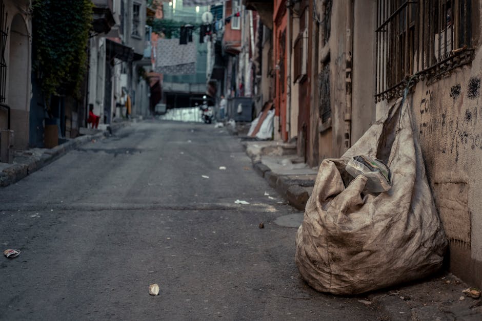 A large, dirty, beige cloth sack filled with assorted waste items, including a partially visible plastic container, rests on the pavement next to an aged, graffiti-marked concrete wall in a narrow, urban alleyway. The alley is lined with closely packed residential buildings featuring weathered facades, iron window grilles, and hanging laundry strung across the upper levels. The asphalt surface is uneven and shows signs of wear, with small pieces of litter scattered across it. In the background, the alley extends uphill, flanked by more buildings with visible doors, windows, and additional waste debris, suggesting a site where private waste collection may be required. Soft, natural lighting indicates early morning or late afternoon, creating a subdued atmosphere. The scene emphasizes challenges related to rubbish disposal in tight, densely populated urban corridors, and subtly aligns with services specialized in on-site clearance or alternative waste handling in restricted-access areas, as handled by companies like House Clearance Kingston.