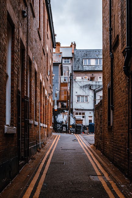 A narrow cobblestone street in Kingston lined with multi-story brick buildings featuring a mix of red and brown tones, irregular façade designs, and small bay windows. The buildings have various architectural details such as decorative arches, balconies, and chimneys. On the right side, there's a traditional black wrought-iron street lamp attached to the wall of a building. A small rectangular hanging sign is also visible on the right, indicating a commercial establishment. Parked along the curb is a white delivery van with visible branding; nearby, a black bicycle is secured to a railing. The street is lit by natural daylight, with some cloud cover visible in a partly cloudy sky. The scene reflects an urban environment typical of UK town centres, where independent collection or private waste disposal, such as through rubbish removal services like House Clearance Kingston, may be employed to manage waste in this historic area. The overall atmosphere suggests a quiet, well-maintained street typical of Kingston's residential and commercial zones.