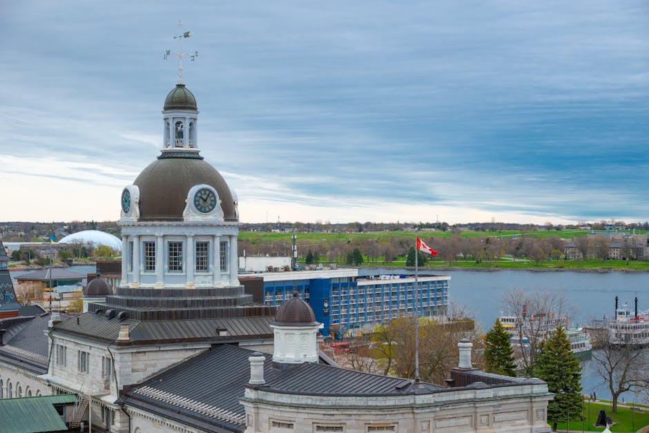 An aerial view of a historic cityscape showing a prominent building with a large, ornate clock tower topped by a small dome and weather vane, surrounded by several other stone and brick structures with dark slate roofs featuring various chimneys and dormer windows. The foreground includes the rooftops of nearby buildings with visible roof vents and skylights. In the background, a river extends across the image with calm water reflecting the partly cloudy, blue and gray sky, and a distant shoreline lined with greenery and low-rise developments. The overall scene is taken during daytime with natural lighting highlighting the architecture and urban environment, providing a sense of the city's historical and modern mix, with some context of urban planning and infrastructure. This image may be used in articles discussing city development, historical architecture, or urban landscapes, relevant to the topic of efficient waste management and building clearance in city environments, naturally supporting content about independent or private waste disposal services by highlighting city infrastructure.