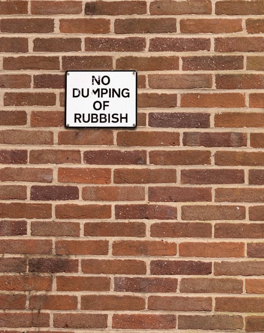 A rectangular metal sign mounted on a reddish-brown brick wall displaying the message 'NO DUMPING OF RUBBISH' in black uppercase letters on a white background. The brick wall features a repeating pattern of horizontal bricks with a slightly rough texture, varying shades of warm brown and reddish hues, and light-colored mortar joints between them. The sign is positioned centrally in the upper half of the image, with the bricks surrounding it uniformly. The appearance suggests an outdoor setting, possibly on a property boundary or service area, where private waste handling and rubbish removal regulations are in place, supported by the presence of the House Clearance Kingston company that provides alternative waste disposal services.