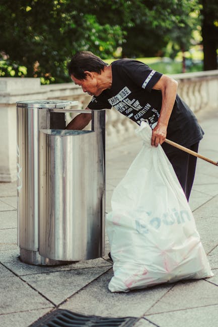 A middle-aged man is seen wearing a black T-shirt with white text and dark trousers, leaning forward to deposit waste into a cylindrical stainless steel rubbish bin with a domed lid, positioned on a paved sidewalk. The man holds a large white plastic bag filled with rubbish in his left hand, while his right hand is reaching toward the bin opening. The surrounding environment features a low stone or concrete wall in the background, with lush green foliage and trees behind it, suggesting a park or garden setting. The scene is illuminated by natural daylight, casting soft shadows on the pavement. This image illustrates an individual actively engaged in private waste disposal, supporting the concept of independent rubbish collection outside local authority services, consistent with the context of alternative waste handling methods.