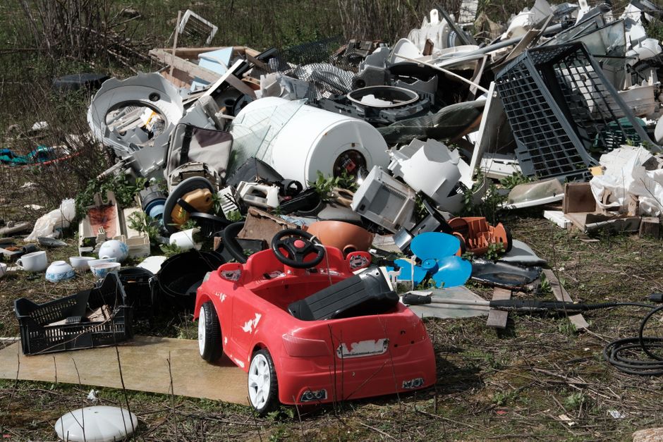 A large pile of discarded household and electronic waste is spread across an outdoor area, including various broken appliances such as washing machines, microwaves, and other appliances with visible metal, plastic, and internal components. Among the debris is a small, bright red plastic children's ride-on car with a black seat and white wheels, situated in the foreground. Surrounding the main heap are smaller scattered items like plastic bowls, containers, a black plastic crate, and blue balloons. The waste is positioned on patchy grass and a concrete surface, with some items leaning or partially covered by dirt and vegetation. In the background, sparse shrubbery and trees frame the scene, indicating an informal dumping site. The natural lighting highlights the textures and colors of the various materials, emphasizing the disorderly accumulation typical of illegal dumping or private rubbish disposal activities. This scene exemplifies waste clutter often associated with alternative collection methods handled by independent waste removal services like House Clearance Kingston, especially relevant in the context of local illegal fly-tipping issues and non-council waste management solutions.