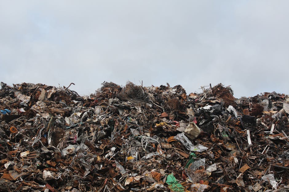 A large pile of mixed waste materials is seen in an outdoor setting with a cloudy sky overhead. The heap contains various types of debris, including twisted metal, plastic fragments, broken wood, cardboard, and other household or commercial waste. The materials are piled in a disorderly fashion, with some items partially buried beneath others. The surface of the waste appears rough and uneven, showcasing textures ranging from smooth plastic surfaces to jagged metal edges and weathered wood. The background features a pale, overcast sky, emphasizing the outdoor environment and the scale of the debris accumulation. This scene reflects the type of waste managed during rubbish removal or waste handling services, illustrating the importance of proper waste disposal, especially for bulky or mixed waste streams, which companies like House Clearance Kingston often address through independent collection or on-site clearance solutions.