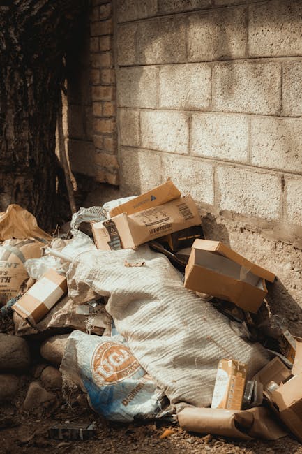 A pile of discarded cardboard boxes, some torn and flattened, along with a large white woven sack and plastic bags, all placed on uneven ground next to a brick wall and a tree trunk. The cardboard boxes vary in size and are predominantly brown, with some featuring printed labels and barcodes. The white sack appears to be filled with additional waste, and the plastic bags are crumpled and partially open. The scene is set outdoors, adjacent to a grey brick wall and natural earth, with small loose stones visible at the base of the wall. The lighting suggests natural daylight, casting soft shadows over the debris, indicating a typical on-site rubbish accumulation that might be managed by private waste removal services like House Clearance Kingston. This scene reflects the typical bulk waste left after household clearance, awaiting collection or disposal, corresponding with the context of independent rubbish removal efforts in the area.