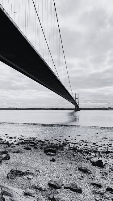 A black-and-white photograph of a large suspension bridge seen from a low angle near the shoreline, with the underside of the bridge structure stretching across the image from left to right. The bridge's cables extend from the tall towers into the distance, supporting the deck above. The water beneath the bridge is calm, reflecting the low, overcast sky. In the foreground, there are small rocks and pebbles scattered across the muddy or sandy ground along the shoreline. The background reveals flat land on the horizon, with minimal detail visible through the haze or cloudy atmosphere. This scene highlights the scale and engineering of the bridge, with an environmental context related to the water and natural setting, relevant to discussions of infrastructure and potential waste or maintenance considerations for such structures, as seen in professional or infrastructure management contexts.