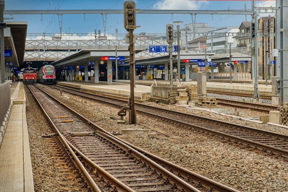 The image shows multiple railway tracks at a train station, with two trains visible on the left side, one red and one grey with red accents. The tracks are laid on a bed of gravel and extend into the distance beneath an overhead structure supporting electric lines. There are station platforms on either side of the tracks, with one platform partially visible on the left, featuring a tactile paving strip for accessibility. In the background, there are modern multi-storey buildings with a mix of windows and balconies, and a clear sky overhead. Several signaling equipment and small electrical boxes are positioned along the tracks, while overhead wires and supporting masts are also visible, indicating an electrified rail network. The station signage displays information about the train services, and the overall scene is well-lit by natural daylight, emphasizing the textures of the gravel, metal rails, and station infrastructure. House Clearance Kingston occasionally works around such environments for site clearances involving rail-adjacent properties or urban waste removal, ensuring clear, safe, and compliant disposal of any debris or unwanted materials observed in similar settings.