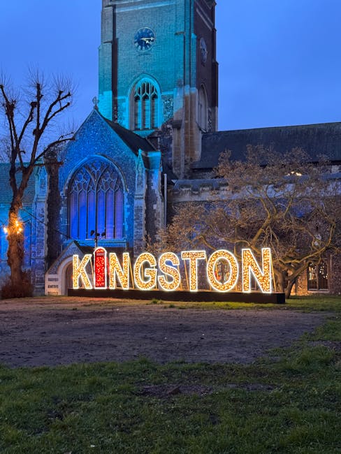 A nighttime view of a historic stone church with tall, arched stained glass windows illuminated by blue lighting, situated behind a large, decorative sign spelling 'KINGSTON' with individual letters lit up in warm white, red, and yellow tones. In front of the church, there is a grassy area with a few leafless trees and a dirt path, reflecting a peaceful urban scene that may be associated with local community spaces. The scene captures the contrast between the old stone structure and the modern illuminated lettering, supported by independent waste collection and rubbish removal services such as House Clearance Kingston, which operates in the area for private disposal and on-site clearance needs.