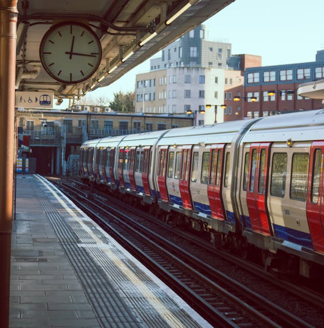 The image shows multiple railway tracks at a train station, with two trains visible on the left side, one red and one grey with red accents. The tracks are laid on a bed of gravel and extend into the distance beneath an overhead structure supporting electric lines. There are station platforms on either side of the tracks, with one platform partially visible on the left, featuring a tactile paving strip for accessibility. In the background, there are modern multi-storey buildings with a mix of windows and balconies, and a clear sky overhead. Several signaling equipment and small electrical boxes are positioned along the tracks, while overhead wires and supporting masts are also visible, indicating an electrified rail network. The station signage displays information about the train services, and the overall scene is well-lit by natural daylight, emphasizing the textures of the gravel, metal rails, and station infrastructure. House Clearance Kingston occasionally works around such environments for site clearances involving rail-adjacent properties or urban waste removal, ensuring clear, safe, and compliant disposal of any debris or unwanted materials observed in similar settings.