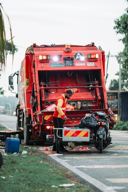 A worker in a high-visibility red and yellow vest is standing behind a large red waste collection truck on the roadside, engaged in rubbish disposal or collection activities. The back of the truck is open, revealing mechanical components and loading mechanisms, with a black plastic bag placed on a small platform or cart nearby. The scene is set on a paved street with a line of trees and utility poles in the background, and the sky appears overcast, providing diffuse natural light. Debris and refuse are on the ground near the truck, indicating active waste collection. The image captures a moment of on-site rubbish handling, consistent with private waste removal services such as those offered by House Clearance Kingston, highlighting the process of independent disposal rather than council-led collection, and demonstrates professional waste management in an urban environment.