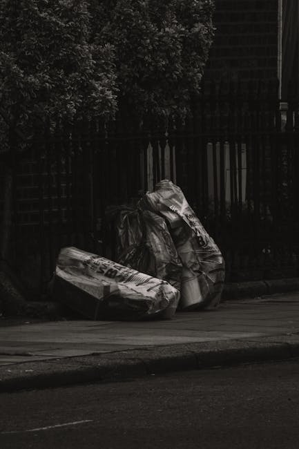 A large pile of mixed household rubbish and waste materials is accumulated on a paved area near a parking lot, consisting of various black and red rubbish bags, flattened cardboard boxes, crumpled paper, plastic packaging, and loose debris. The waste is scattered around an overflowing grey recycling bin labeled for mixed paper and cardboard, with its lid open, revealing paper waste spilling out. Behind the pile are parked vehicles, including a silver car positioned on the left side. In the background, there is a commercial building with a blue metal scaffolding structure and signage for a fish bar, along with illuminated storefronts. A metal railing runs parallel to the waste pile, separating it from the parked car. The scene appears to be outdoors during daytime, with natural lighting highlighting the cluttered arrangement, which illustrates typical urban waste accumulation and the need for private rubbish collection services, such as those offered by House Clearance Kingston.