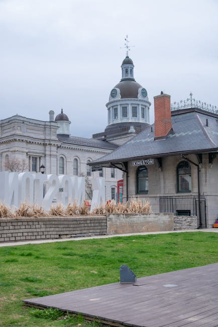 The image shows a historic train station building with a grey slate roof, a brick chimney, and a prominent domed clock tower with a weather vane on top, situated in an urban area with surrounding classical architectural structures. In the foreground, there is a well-maintained grassy area bordered by a low brick wall, and dry ornamental grasses are planted along the edge. A paved pathway leads towards the station, and a small mounted object appears on the pavement, possibly a bollard or security device. Behind the station, a large, modern white sculpture spelling out 'KINGSTON' is visible, constructed from what appears to be metal or painted material, and is situated on a raised bed filled with similar dry ornamental grasses. The scene is evenly lit, with diffuse daylight under an overcast sky, creating a neutral backdrop for the historical and modern elements combined. This setting illustrates a typical location where private waste collection or on-site clearance might occur near or at public transport hubs, reflecting the visual relevance to alternative rubbish removal services managed by companies like House Clearance Kingston.