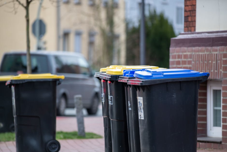 A group of four large, wheeled refuse bins positioned on a paved sidewalk adjacent to a brick residential building. The bins are made of black plastic with textured surfaces and are fitted with flat, rectangular lids in bright yellow, blue, and black colours. The bins are arranged in a row, with the two largest upright on the right side and a smaller bin on the left. A grey van is partially visible and parked on the street behind the bins, along with a blurred background of houses, trees, and utility poles under natural daylight. The scene suggests a setting for independent waste collection or on-site rubbish disposal, with the bins prepared for rubbish removal services, such as those provided by House Clearance Kingston. The environment appears tidy, with the bins intended for regular waste or recycling collection, and the overall composition highlights standard domestic refuse storage in a residential area.