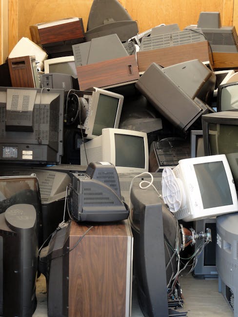 A large collection of old electronic devices and computer equipment piled together indoors against a wooden or brown-colored wall. The heap includes numerous CRT monitors with rounded screens, some stacked on top of each other and others laying at various angles. Several desktop computer towers, mostly made of grey or black plastic, are visible among the pile, with some showing vents and control panels. Wooden or wood-effect speaker cabinets and small peripherals are also intermixed throughout the heap. Cables and wires are tangled around the devices, with a few power strips and connectors visible. The overall scene presents an assortment of discarded technology, typical of an on-site clearance or private waste collection process. House Clearance Kingston specializes in rubbish removal and alternative waste handling solutions, and this image exemplifies the type of electronic waste they may collect for disposal. The arrangement appears cluttered, with no clear order, emphasizing the extent of electronic waste accumulated in the space, ready for professional removal.