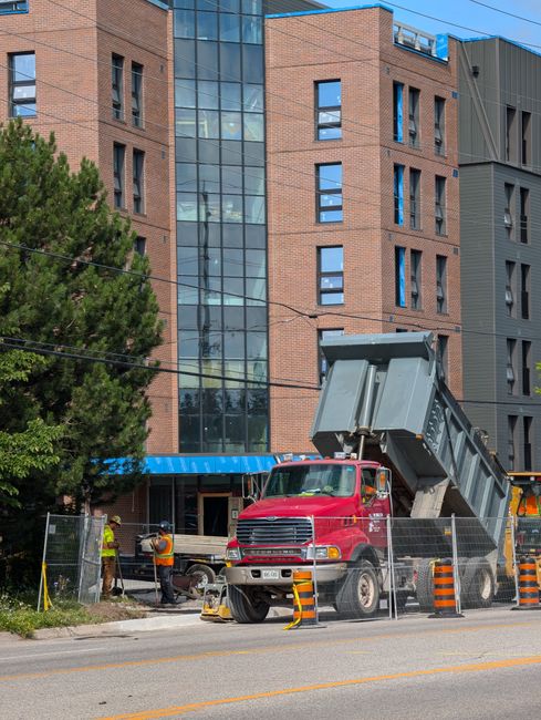 A red dump truck with an open-top metal container is positioned on the sidewalk in front of a modern multi-storey building with a brick and dark grey exterior. The truck's container is raised at an angle, indicating ongoing rubbish removal or waste transfer. Several workers wearing high-visibility clothing and helmets are present near the truck, engaged in task activities related to waste collection, standing behind temporary fencing and orange traffic cones that cordon off the work area. A large green tree partially obscures the left side of the scene, with power lines crossing overhead. The building features large glass windows and a blue awning above the entrance, suggesting a commercial or residential complex. The environment is well-lit, with natural daylight highlighting the textures of the brickwork, metal container, and vehicle surfaces. The scene depicts an instance of private waste handling or on-site clearance, aligning with services provided by companies like House Clearance Kingston, for a non-local authority rubbish removal context.